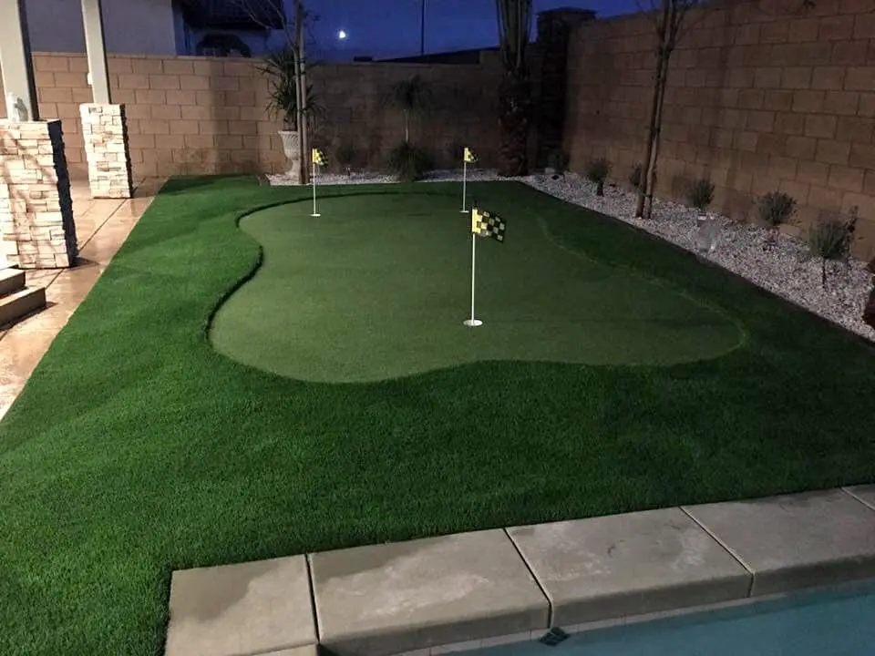 A backyard scene with a well-manicured lawn surrounded by a brick patio. An orange tree bearing ripe oranges stands to the left, and large pine trees and a wheelbarrow are in the background. Sunlight casts shadows over the synthetic turf, creating a peaceful ambiance.