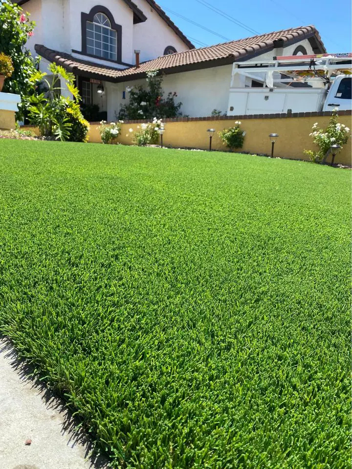 A lush, well-manicured green lawn stretches across the foreground, bordered by a stone path. Featuring custom putting green installations with artificial turf, it offers pet-friendly lawns for every family member. In the background, a house with a tan exterior and tiled roof stands beside a parked white truck.