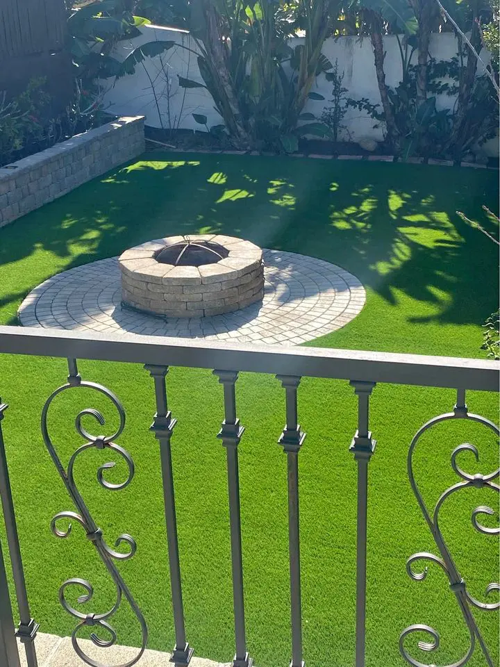 A backyard features a circular stone fire pit on a tiled area, surrounded by synthetic turf for easy maintenance. Decorative metal railings grace the foreground, while lush plants line the back wall, offering a stunning landscape enhancement.