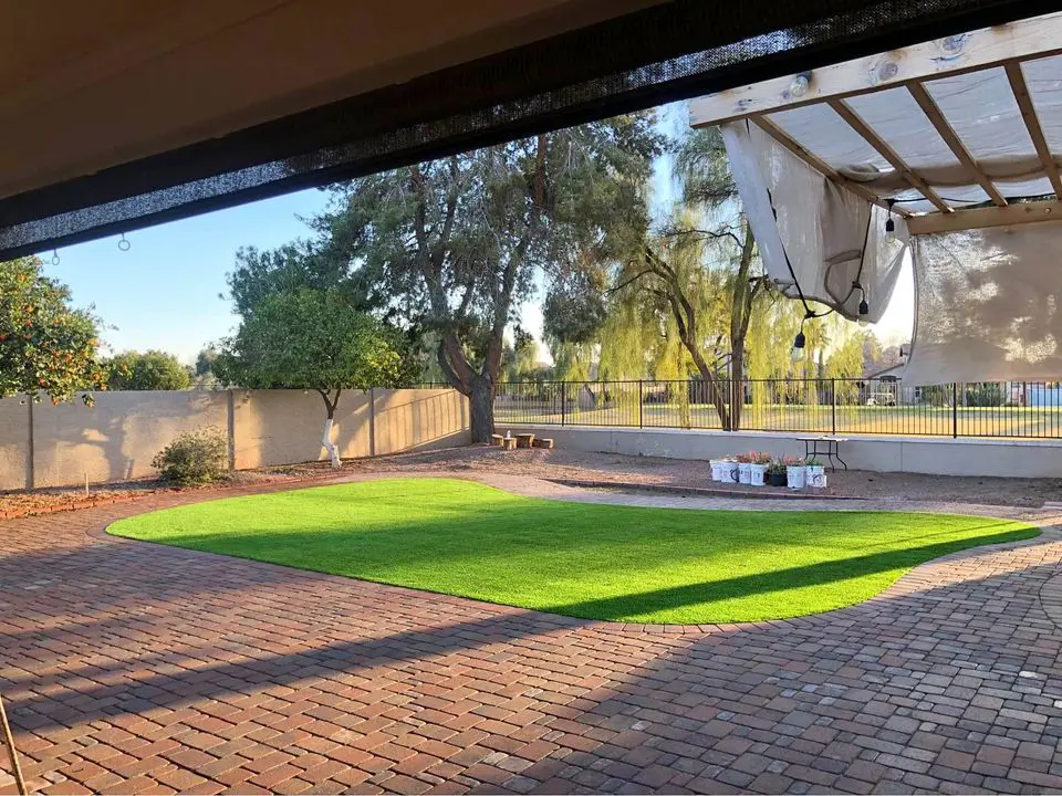 Backyard with a section of artificial turf surrounded by a brick patio. Trees and a low wall border the yard. A pergola with hanging sheets provides partial shade. Gardening supplies and plants sit in a corner, suggesting the work of skilled artificial grass installers. Clear blue sky in the background.