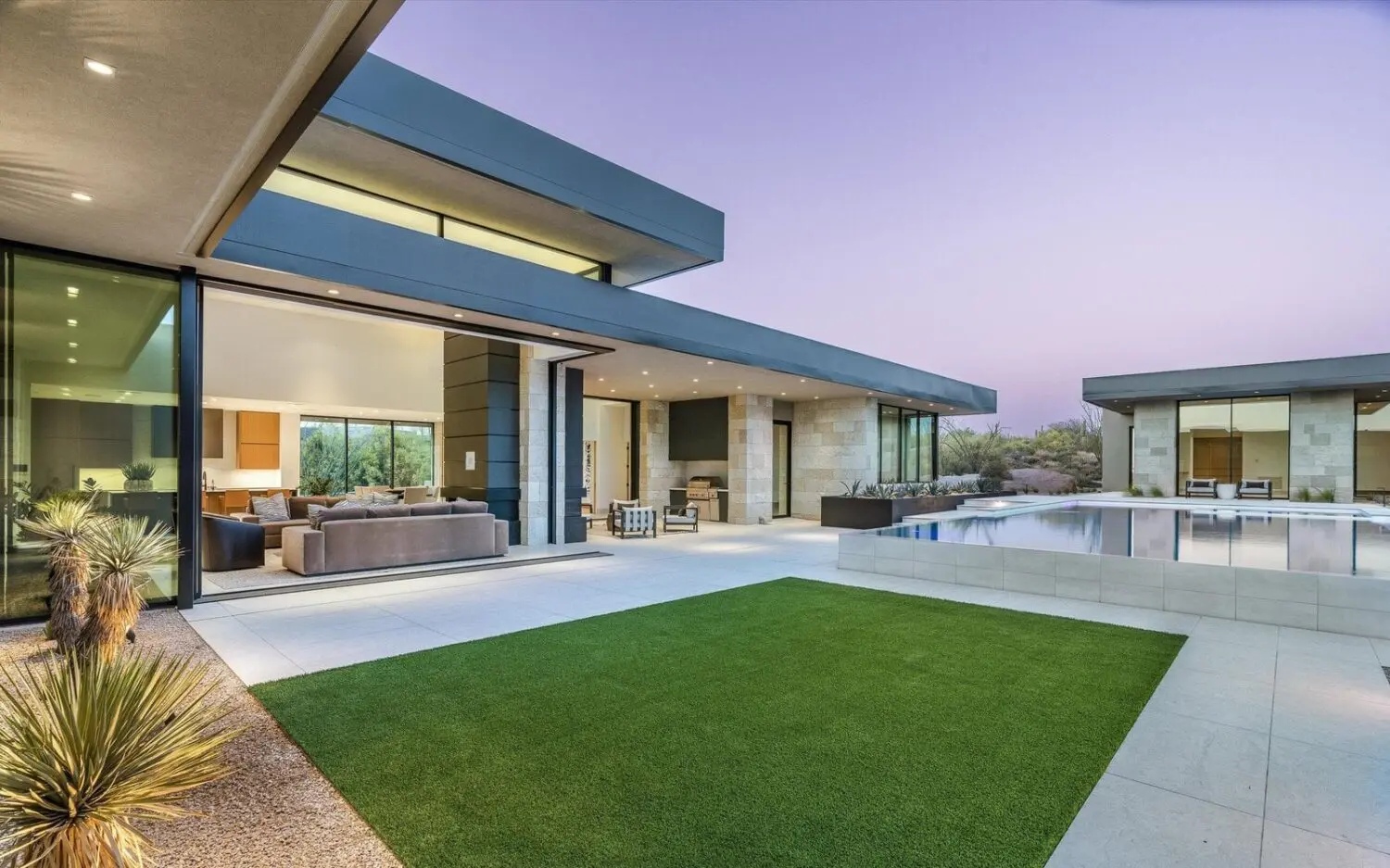 Modern home exterior with large windows, geometric design, and a blend of stone and metal finishes. The patio features a lush, rectangular section of synthetic turf and a pool reflecting the twilight sky. Desert plants line the gravel path, offering natural landscape enhancement.