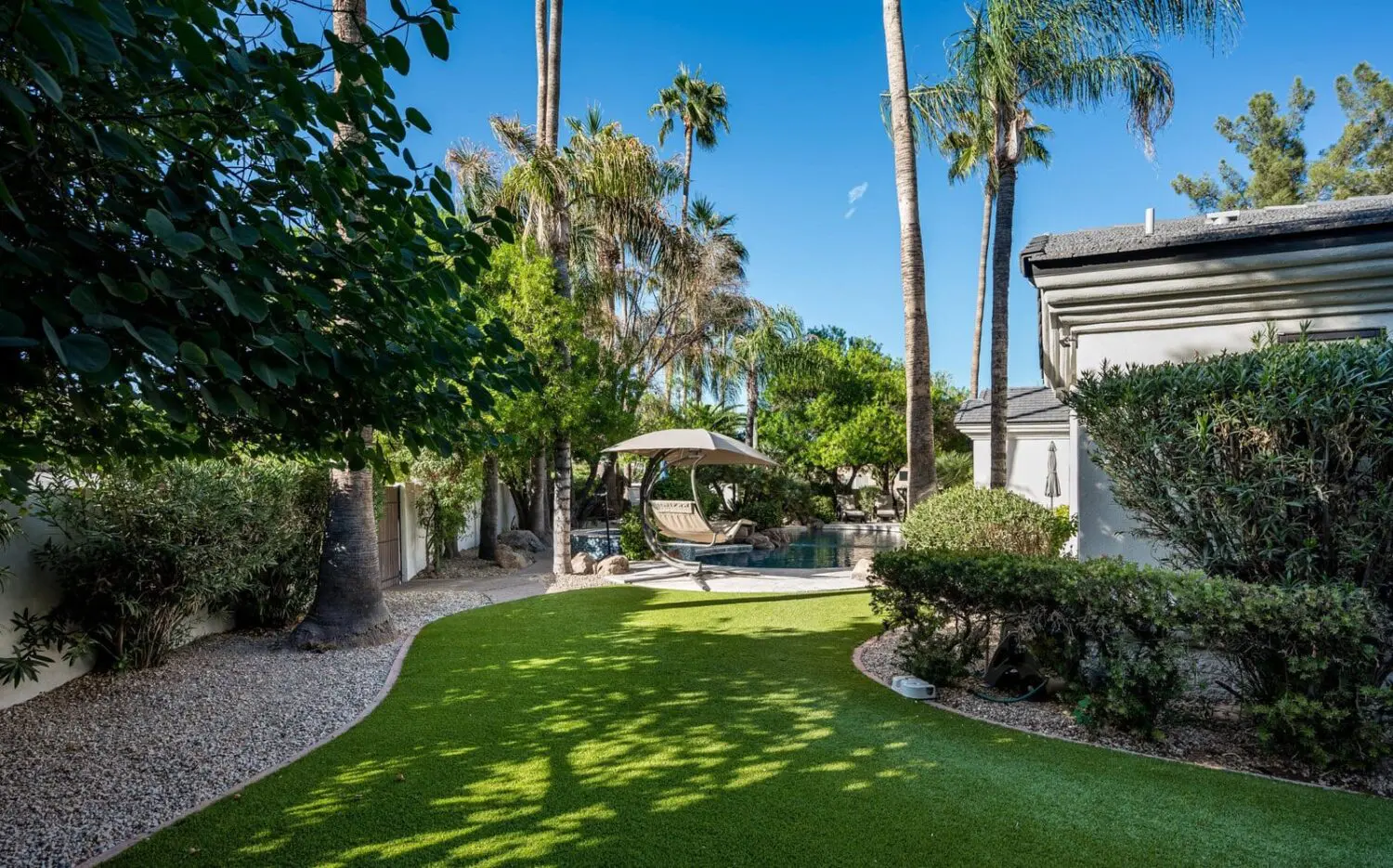 A lush backyard featuring a curved pathway of vibrant artificial turf, surrounded by palm and leafy trees. In the background, a hammock rests beneath a shaded structure near a small pool, with the tranquil blue sky overhead—a paradise made easy with expert artificial grass installers.