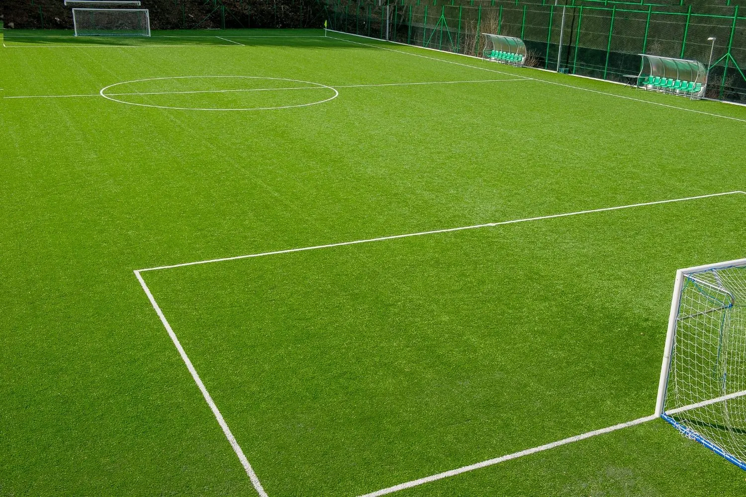 The empty soccer field boasts bright green synthetic turf, with goals at both ends and crisp white markings outlining boundaries and the center circle. Benches are alongside, while a netted fence runs the perimeter. Artificial grass installers ensure quality despite installation costs.