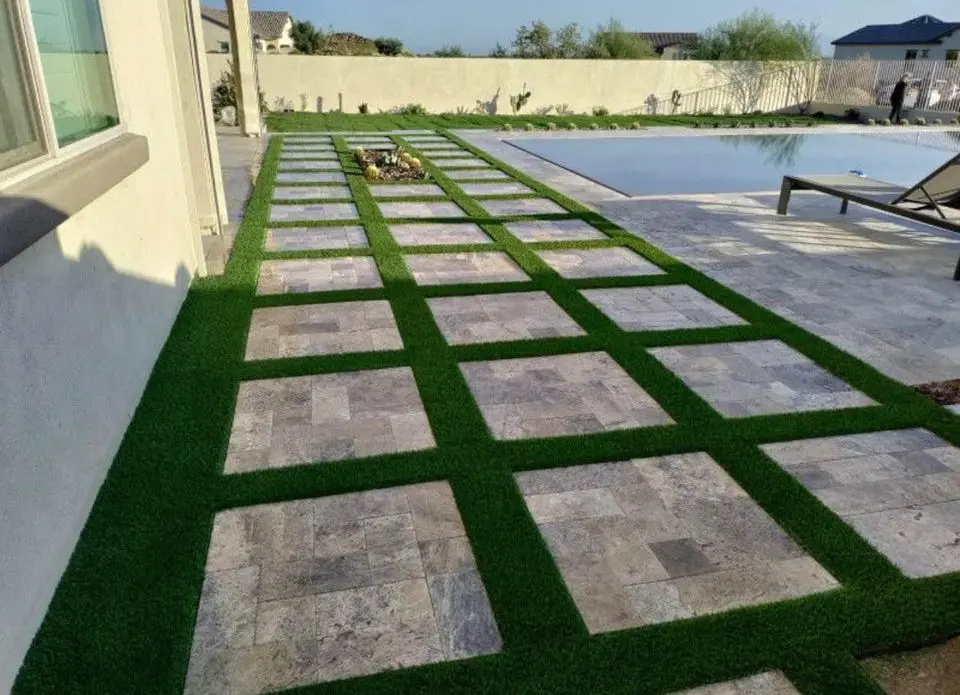 Modern backyard with a stone-tile pathway interspersed with synthetic turf, leading to a rectangular pool. A lounge chair rests on a stone patio near the pool. In the background, a low wall and plants are visible under a clear blue sky.