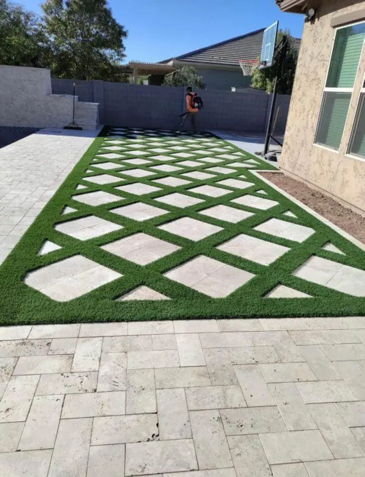 In Sun City, AZ, a backyard showcases a modern design with synthetic turf and grass in a checkerboard pattern. A person uses a leaf blower near the basketball hoop, while the sun casts its glow over the concrete fence and house exterior.