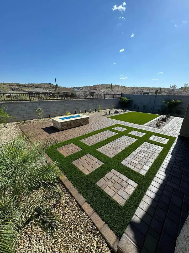 A backyard showcases a geometric pattern of pavers and artificial grass, laid by expert artificial grass installers, with a stone outdoor fireplace. A gravel path lined with various plants borders the yard, while a brick wall and desert landscape bask under the clear blue sky.