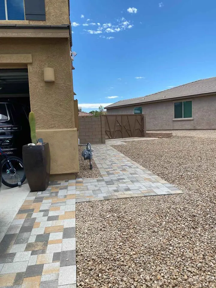 A house with a tan exterior features a paved driveway on the left and a gravel front yard, designed with pet-friendly lawns. A metal bird sculpture stands near the corner, while a large ceramic planter with a cactus sits by the entrance. Another house is visible in the background.