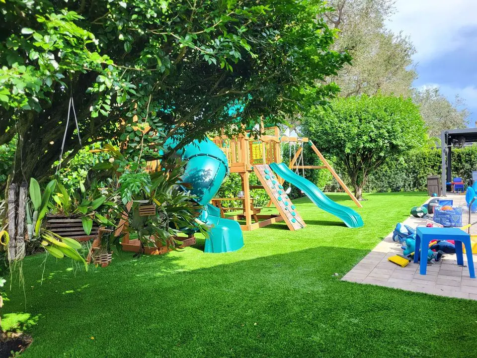 A backyard playground with a green slide, climbing wall, and rope is surrounded by lush greenery and trees. Neatly trimmed artificial turf covers the ground, adding a fresh touch. There's a paved area with blue chairs and scattered toys. It's a bright, sunny day with clear skies.