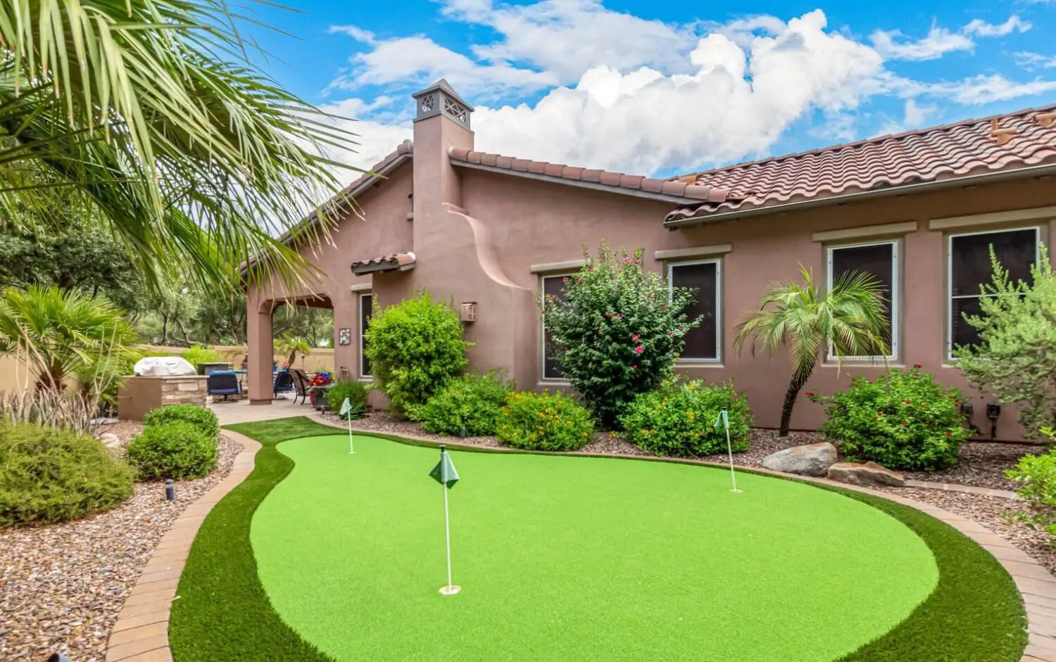 A backyard in Goodyear, AZ features a small putting green crafted from high-quality artificial turf, surrounded by well-maintained shrubs, palm trees, and stone landscaping. A stucco house with a tiled roof stands in the background under a blue sky with a few clouds.