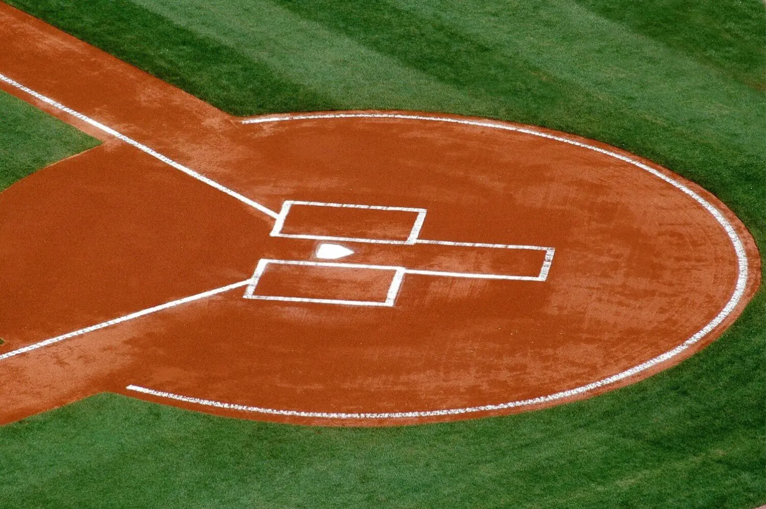 Aerial view of a baseball field's home plate area. The dirt is neatly raked with white chalk outlining the batter's boxes and base lines. Synthetic turf surrounds the clay, its lush green contrasting beautifully with the red-brown surface, showcasing expert craftsmanship by artificial grass installers.