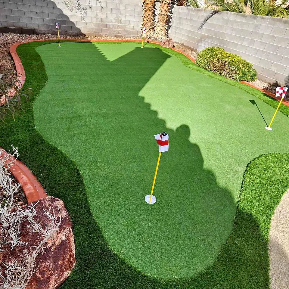 A small artificial putting green, showcasing pristine turf in a pet-friendly backyard, is bordered by rocks and plants with three golf flags. A shadow is cast over part of the green, while a brick wall and palm tree create a serene backdrop.