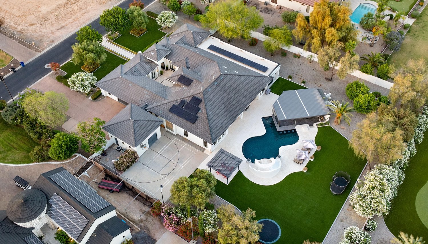 Aerial view of a residential property with a large house, swimming pool, basketball court, and well-maintained synthetic turf lawn. The property is surrounded by trees and shrubs, with neighboring houses visible nearby.