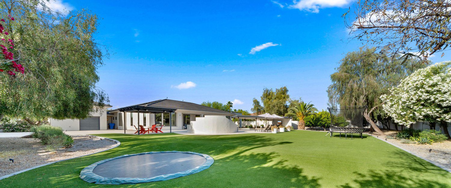 A spacious backyard featuring a green lawn with an in-ground trampoline, shaded seating area with umbrellas, and a modern house with large windows in the background. With synthetic turf installations enhancing its beauty, the yard is surrounded by trees and bushes under a clear blue sky.