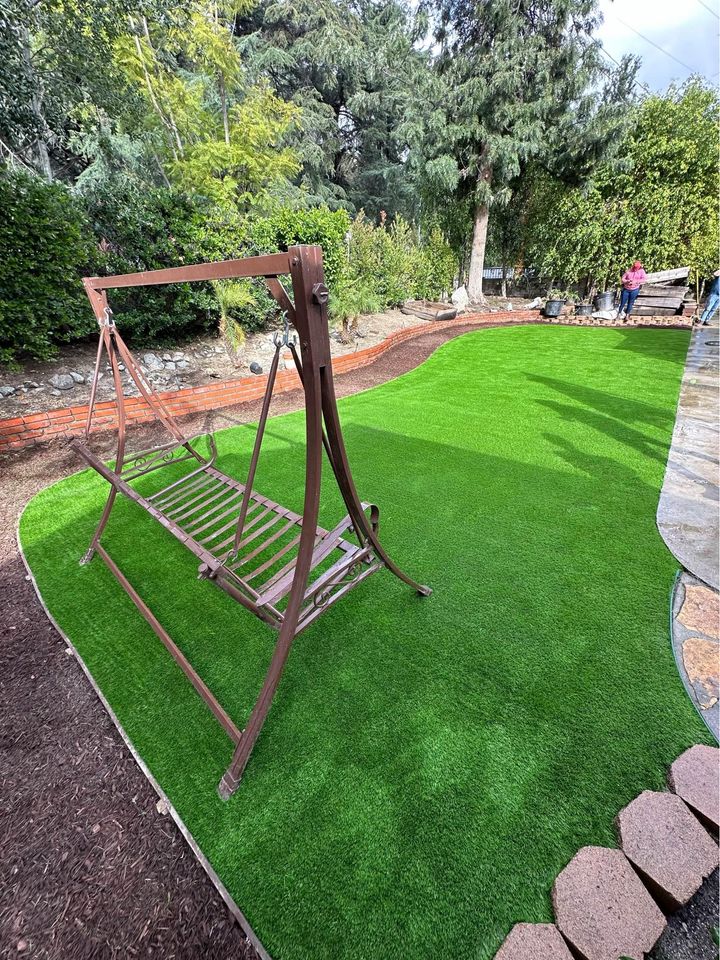 A garden in Surprise, AZ features a metal swing frame without the swing, set on a neatly manicured, lush green lawn. Surrounding the artificial turf are various trees and bushes, with a person and a paved walkway visible in the background.