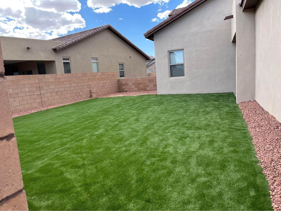 A backyard scene with lush green synthetic turf surrounded by beige stone gravel. The area is enclosed by a high brick wall, and the backyard is bordered by two beige stucco houses. The sky above is blue with scattered clouds.