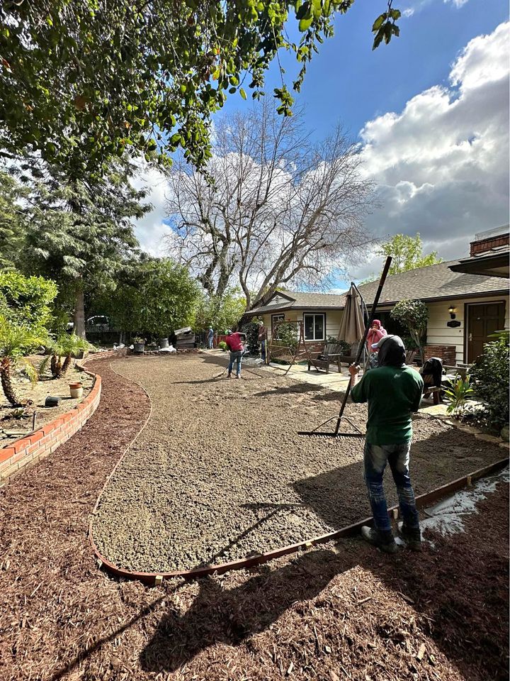 A backyard scene with several people spreading gravel across a large area using tools, preparing for a synthetic turf installation. A tree without leaves stands near the center. The sky is partly cloudy, and the surrounding garden is lush with greenery, a small brick wall, and a house in the background.