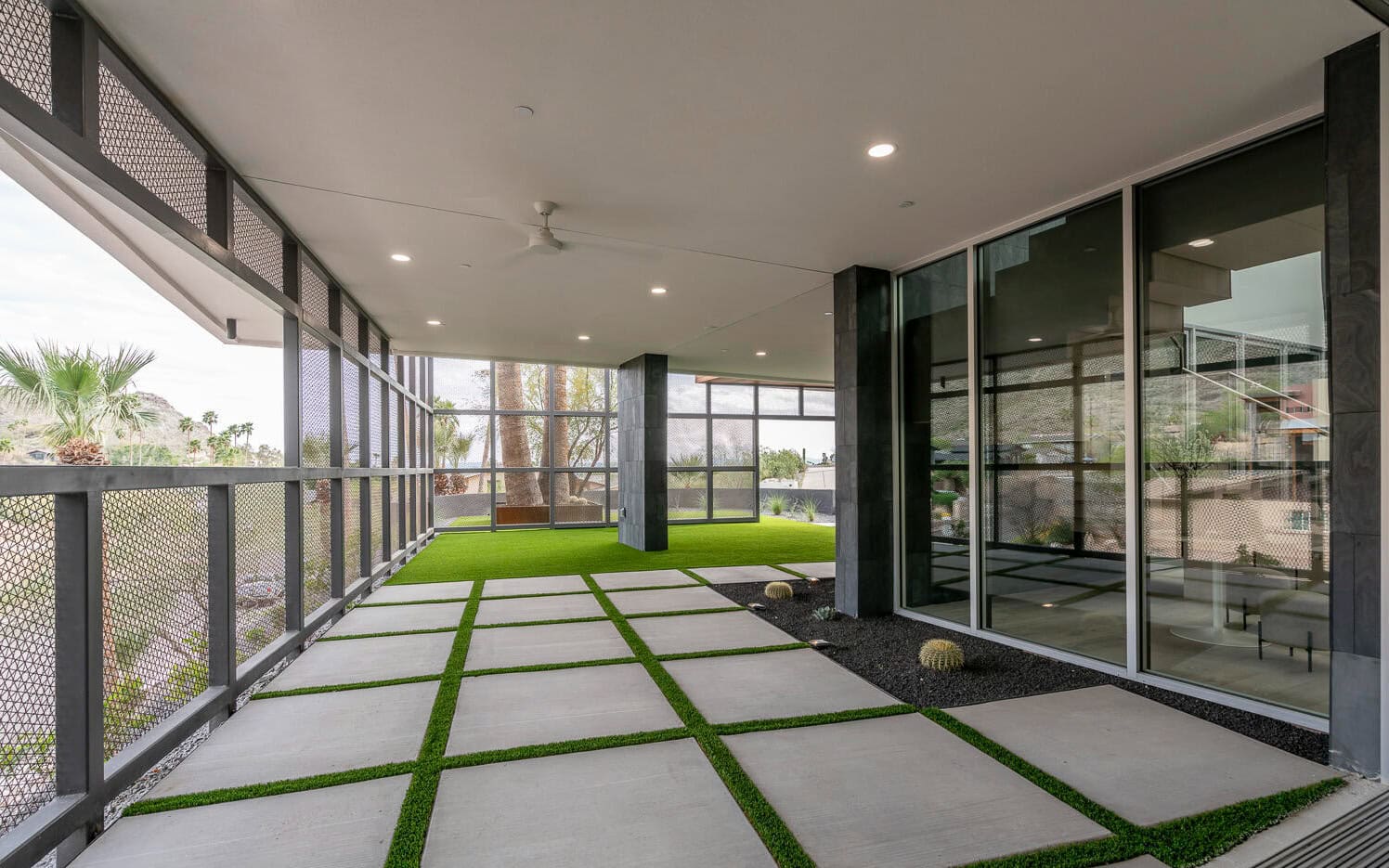 A modern patio in Surprise, AZ, with large glass windows, features a tiled floor interspersed with synthetic turf. The area is enclosed by mesh walls for partial outdoor views. A ceiling fan is installed above, and desert plants are visible outside.