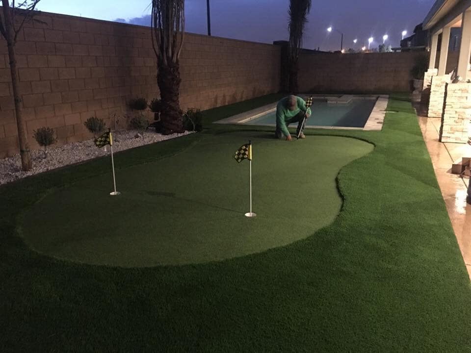 A person kneels next to a small artificial putting green, a testament to the skill of an artificial turf installer, with two flags in a backyard. Beside the putting green is a narrow rectangular swimming pool. The area is bordered by a brown brick wall and features some landscaping with small plants and rocks.