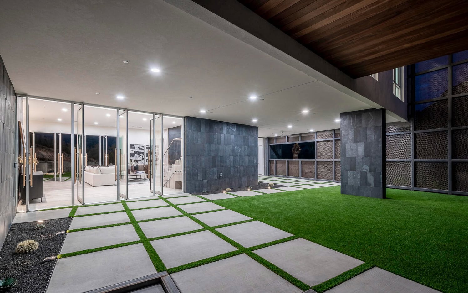 A modern outdoor space featuring grassy areas and sleek paver installations intersected by large square concrete tiles. The courtyard, with its contemporary design, leads to an interior living area visible through floor-to-ceiling glass walls. The space is illuminated by recessed lighting.