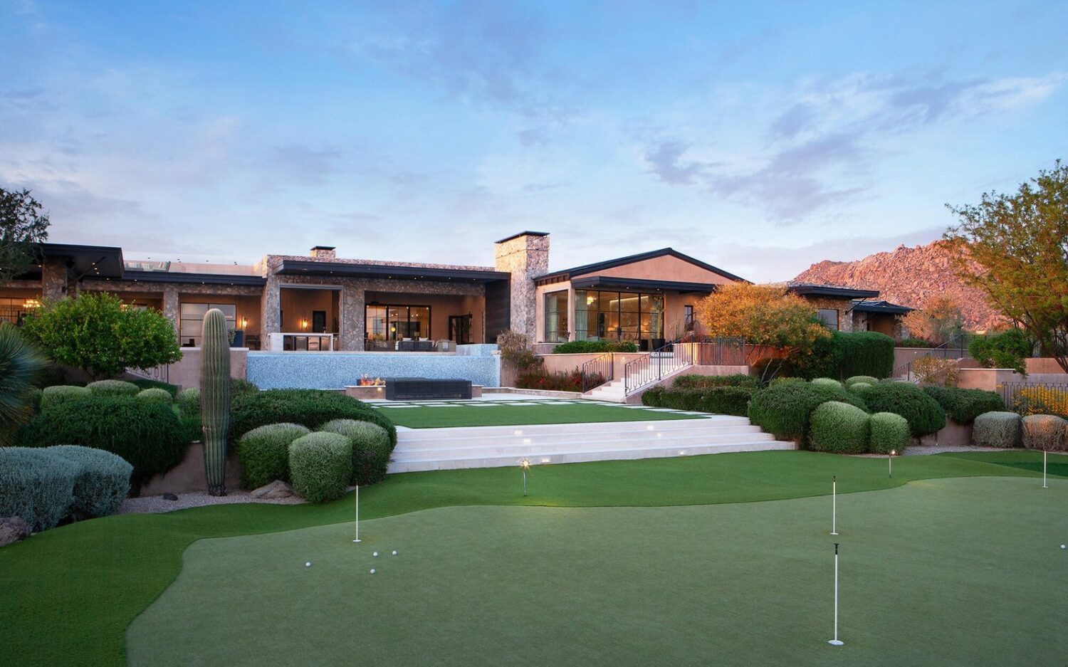 A luxurious desert home in Surprise, AZ, with large windows, modern architecture, and a spacious outdoor area that includes a putting green made of synthetic turf. The landscape features manicured lawns, desert plants, and a backdrop of mountains against a twilight sky.